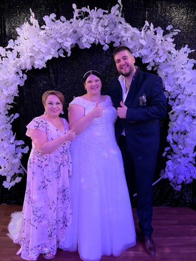 Drei Personen posieren vor einer Blumendekoration bei einer Feier. Die Braut trägt ein weißes Kleid.