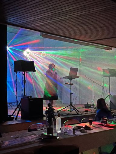 Eine Person singt auf einer Bühne, umgeben von bunten Lichtstrahlen und Nebel.