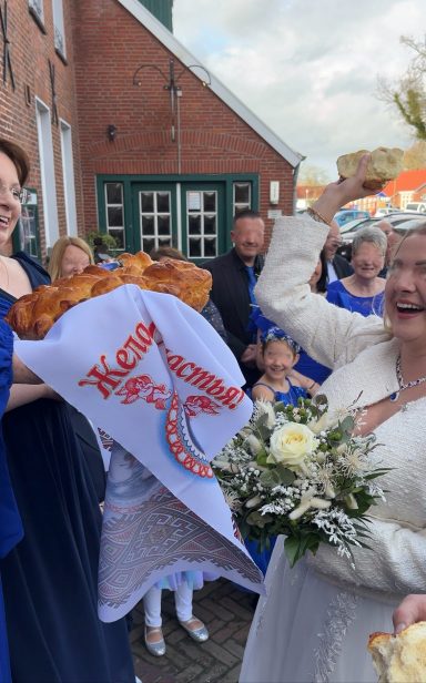 Eine Frau in Weiß hält einen Blumenstrauß und jubelt, umgeben von feiernden Gästen.