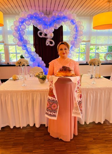 Frau in rosa Tracht mit Brot vor festlich dekoriertem Tisch und Herz aus Ballons.