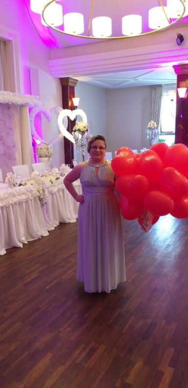 Frau in grauem Kleid hält viele rote Luftballons in einem festlich dekorierten Raum.