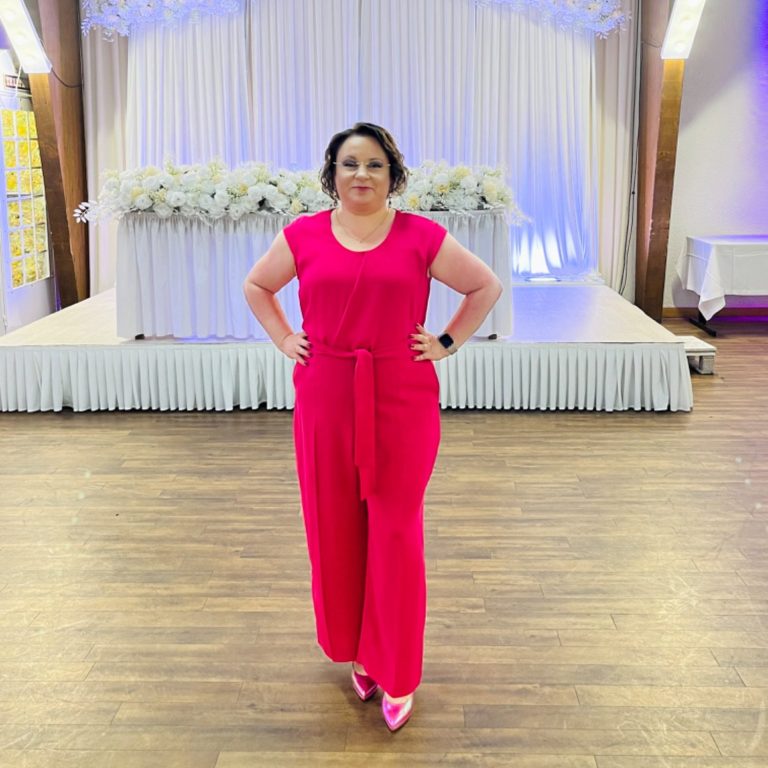 Frau in pinkem Jumpsuit mit Hand an der Hüfte, steht vor einer festlichen Kulisse.