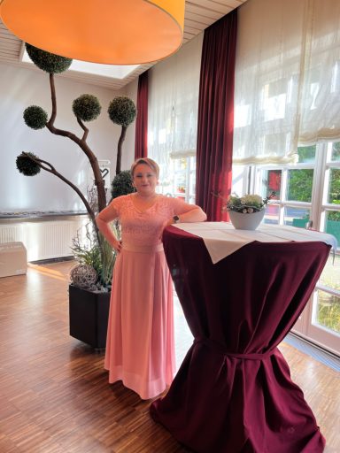 Frau in einem rosa Kleid steht neben einem Tisch in einem stilvollen Raum.