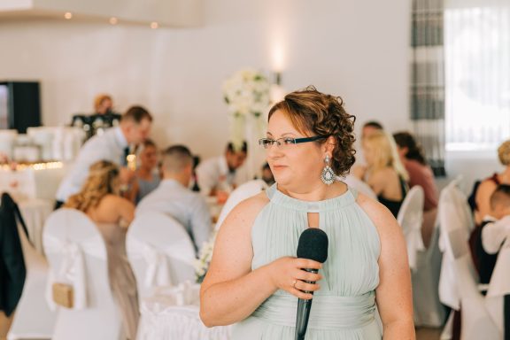 Eine Frau in einem hellen Kleid hält ein Mikrofon, während Gäste an einem Tisch sitzen.