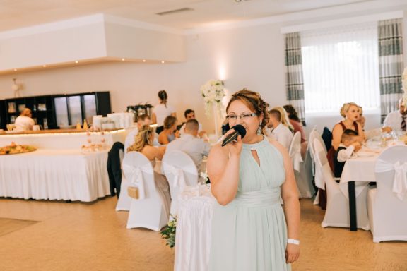 Frau mit Mikrofon in einem eleganten Veranstaltungsraum, Gäste im Hintergrund.