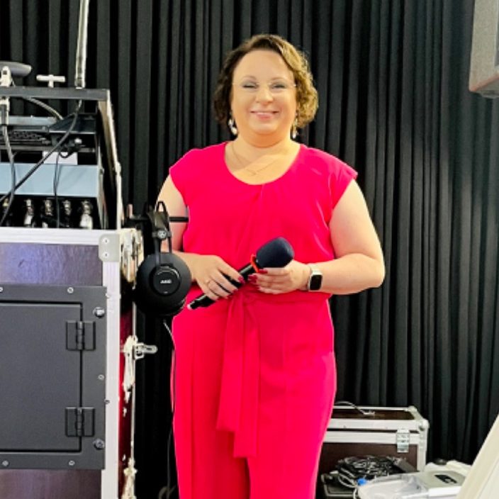 Frau in pinkem Outfit hält ein Mikrofon, lächelt vor schwarzem Vorhang.